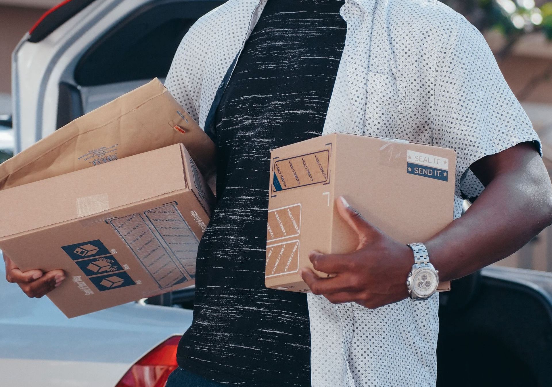 A cheerful man holding packages stands by an open car trunk outdoors, ideal for delivery and logistics concepts.